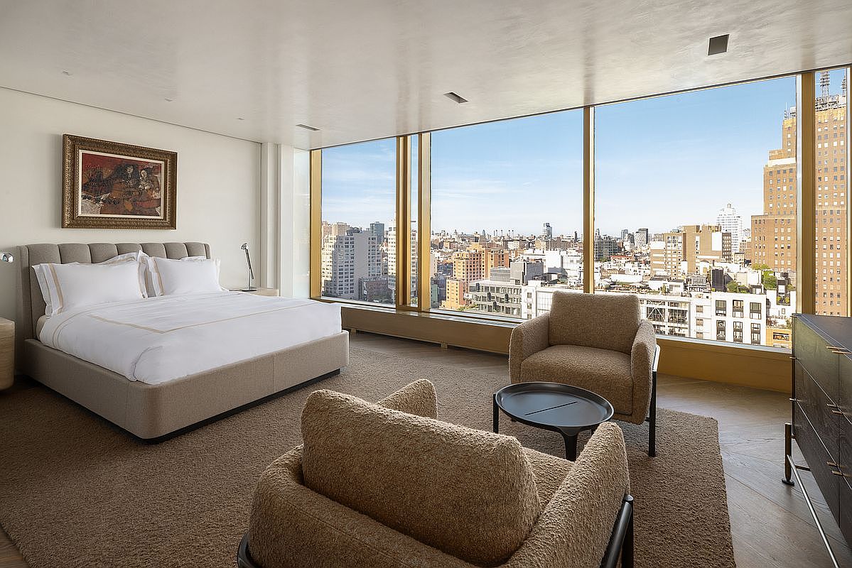 This primary bedroom features a neutral color palette and a plush, upholstered bed frame centered against a wall adorned with a framed piece of art. The room is defined by expansive floor-to-ceiling windows that provide a panoramic view of the city skyline, bathing the space in natural light. A cozy seating area with two textured armchairs and a small circular table creates a comfortable lounge space, while the overall aesthetic is sophisticated and minimalist.