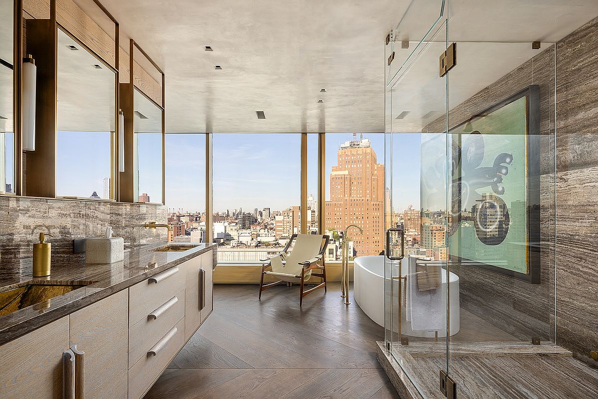 This luxurious primary bathroom features a sleek, modern design with a floating light wood vanity, stone countertops, and a glass-enclosed shower. A freestanding soaking tub is positioned near expansive floor-to-ceiling windows that offer a dramatic, panoramic view of the city skyline. The room is finished with rich, patterned wood flooring and warm, ambient lighting, creating a sophisticated and serene atmosphere.