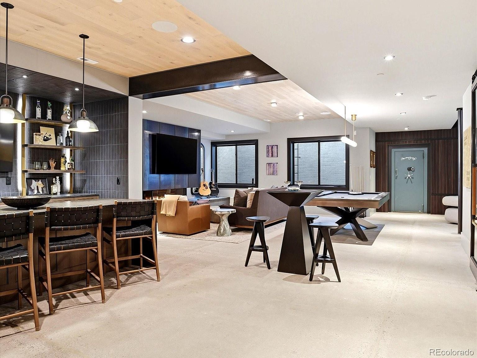 This expansive, modern basement features a sophisticated entertainment area complete with a wet bar, a cozy living space with a fireplace, and a dedicated billiards zone. The design incorporates a mix of industrial and warm elements, including concrete flooring, wood-paneled ceilings, and dark, moody accent walls. The open-concept layout creates a seamless flow, making it an ideal space for hosting and relaxation.