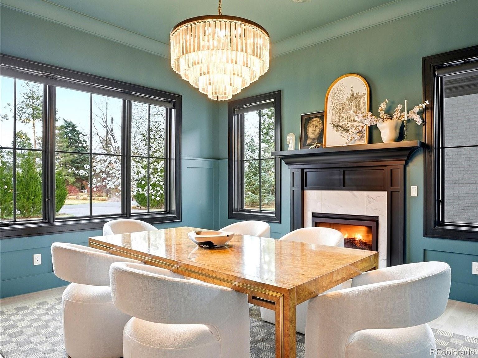 This elegant dining room features a sophisticated teal color palette with dark trim, centered around a polished burl wood table and six plush, cream-colored curved chairs. A striking tiered crystal chandelier hangs above the table, while a cozy fireplace with a marble surround and curated decor serves as a focal point. Large windows with black frames provide ample natural light and a view of the lush outdoor greenery, creating a refined and inviting atmosphere.
