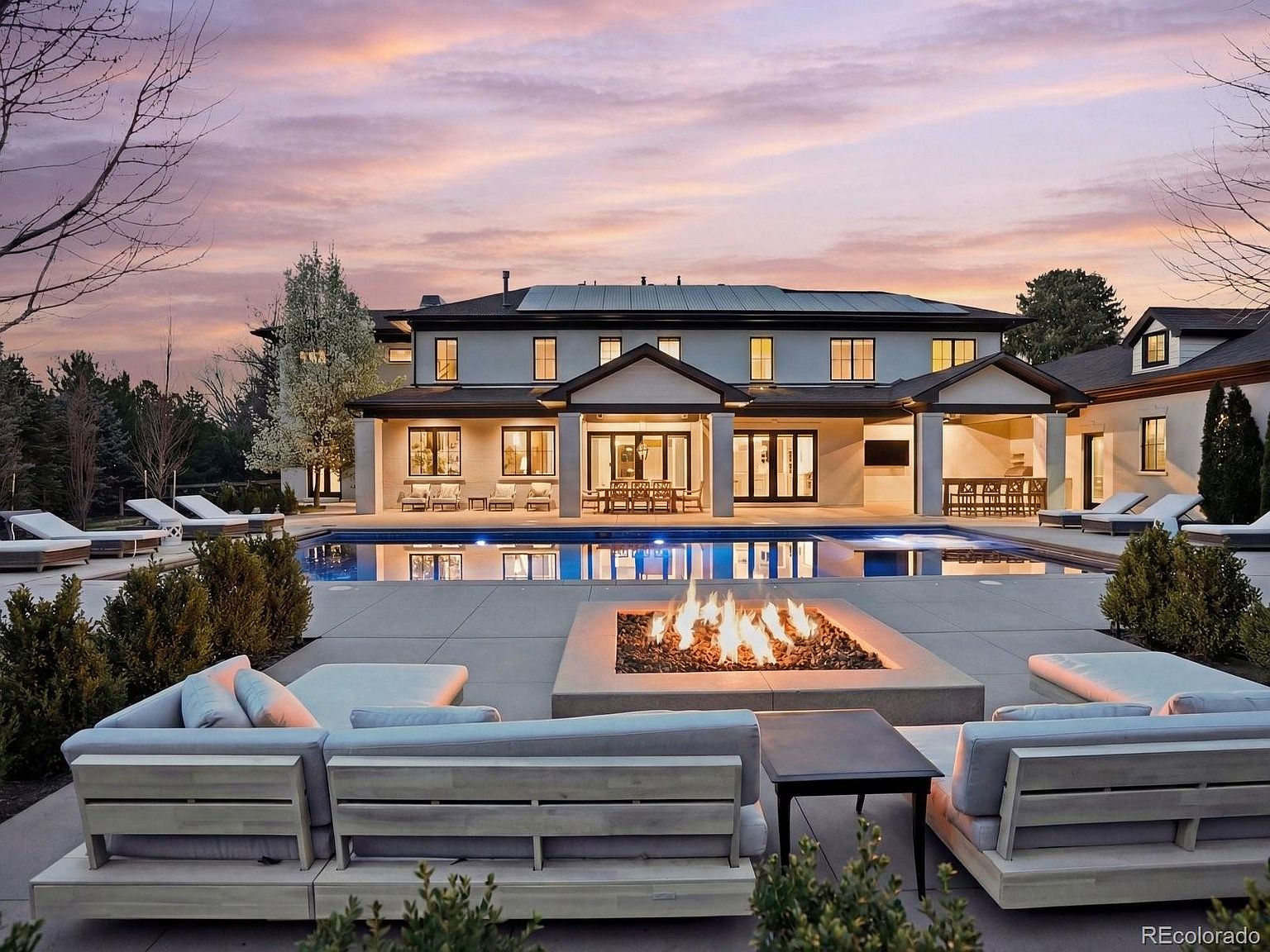 This stunning rear exterior view showcases a luxurious modern estate at sunset, featuring a sprawling patio with a central fire pit and a large, pristine swimming pool. The home boasts a symmetrical architectural design with clean lines, multiple windows, and a covered outdoor living area perfect for entertaining. The warm, golden-hour lighting enhances the inviting atmosphere, highlighting the seamless transition between the indoor and outdoor living spaces.