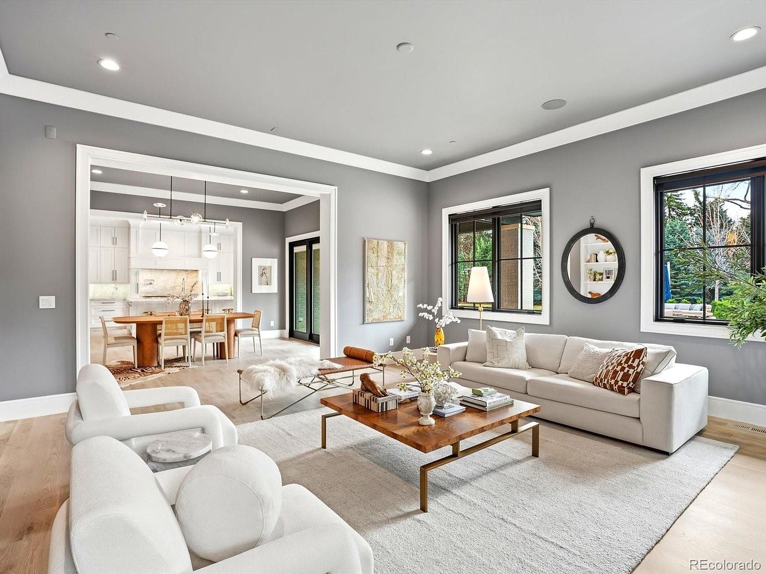 This bright and airy living room features a sophisticated, neutral color palette with plush white seating, a warm wooden coffee table, and a textured area rug. The open-concept design seamlessly flows into an adjacent dining area, creating a spacious and inviting atmosphere perfect for entertaining. Large windows with dark frames provide ample natural light and a view of the outdoors, while the clean lines and modern decor emphasize a contemporary, high-end aesthetic.
