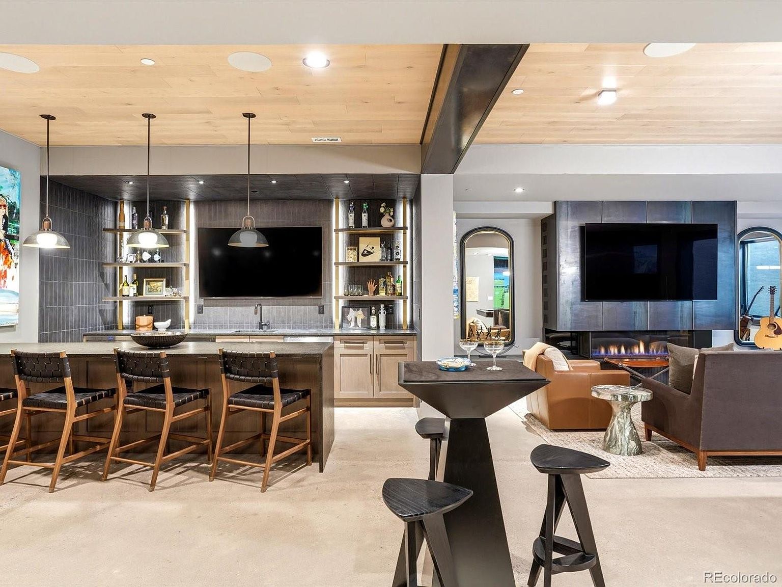 This sophisticated basement living area features a sleek, modern wet bar with dark cabinetry, open shelving, and three woven bar stools. Adjacent to the bar is a cozy lounge space centered around a contemporary fireplace with a large wall-mounted television and comfortable seating. The room is finished with a light wood-paneled ceiling and polished concrete floors, creating a luxurious and inviting entertainment space.