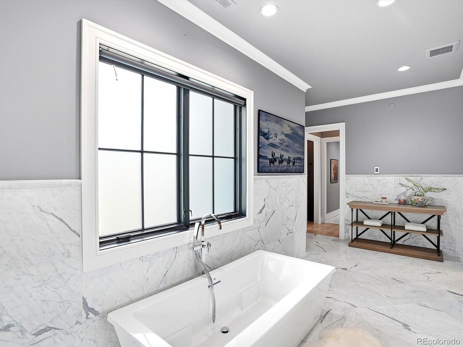 This luxurious primary bathroom features a sleek, freestanding white soaking tub positioned beneath a large, black-framed window. The walls are adorned with elegant white marble wainscoting that complements the matching marble flooring, creating a sophisticated and serene atmosphere. A rustic wooden console table adds a touch of warmth to the space, while a framed landscape print enhances the refined aesthetic.