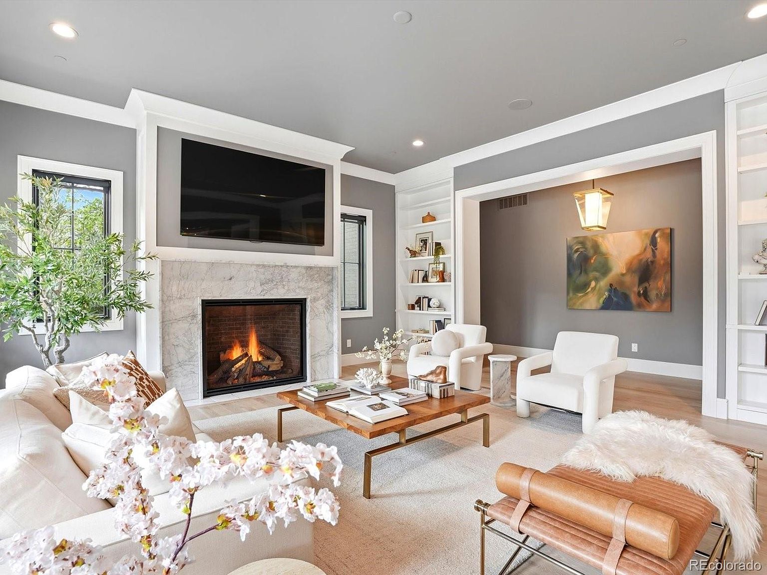 This elegant living room features a sophisticated neutral color palette with a striking marble-faced fireplace as the central focal point, topped by a large wall-mounted television. The space is furnished with plush cream-colored seating, a warm wooden coffee table, and a leather bench, all arranged on a soft area rug. Built-in shelving units flank the fireplace and the adjacent alcove, which is highlighted by a modern pendant light and a large piece of abstract wall art, creating a luxurious and inviting atmosphere.