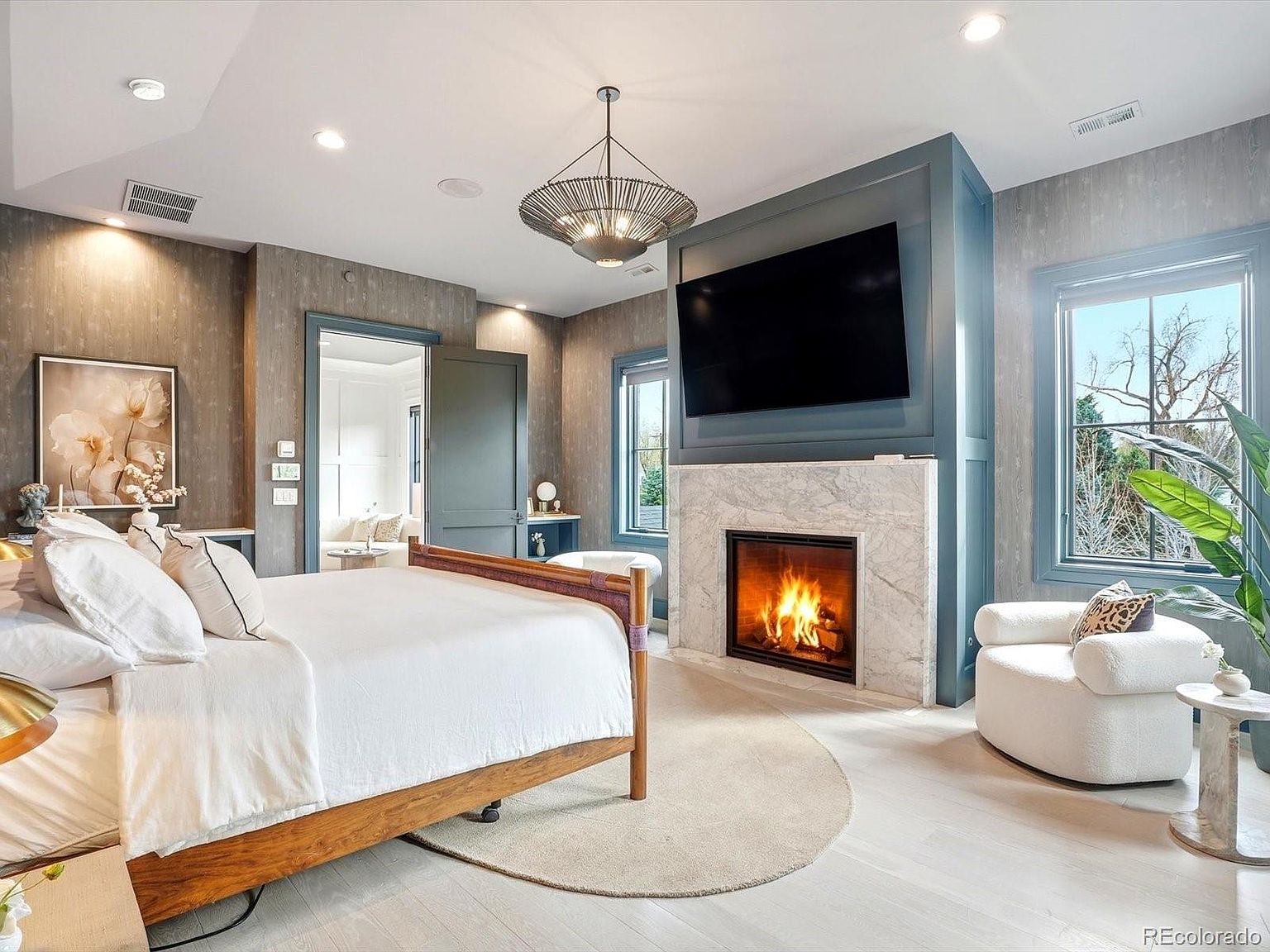 This elegant primary bedroom features a warm wooden bed frame, a cozy white armchair, and a striking fireplace with a marble surround topped by a large wall-mounted television. The room is accented with textured wallpaper, a modern chandelier, and light-toned flooring, creating a sophisticated and tranquil atmosphere. The perspective is a wide-angle shot from the foot of the bed, capturing the inviting layout and the seamless integration of comfort and luxury.