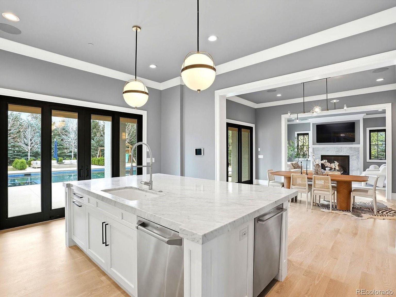 This bright and airy kitchen features a large white island with a marble countertop, stainless steel appliances, and elegant globe pendant lighting. The open-concept design seamlessly transitions into a formal dining area, with large glass doors providing a view of the outdoor pool area. The space is defined by its clean lines, neutral gray walls, and light hardwood flooring, creating a sophisticated and inviting atmosphere.