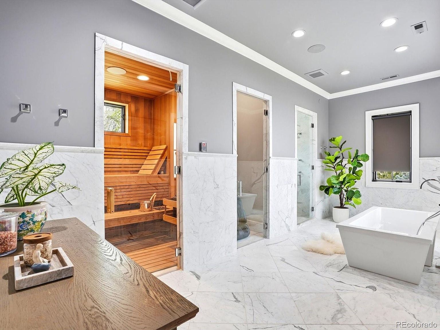 This luxurious primary bathroom features a high-end sauna with wood paneling, a sleek freestanding soaking tub, and a separate glass-enclosed shower area. The space is finished with elegant white marble flooring and wainscoting, complemented by a large potted plant and a wooden vanity surface. The bright, airy atmosphere and premium materials create a spa-like retreat within the home.