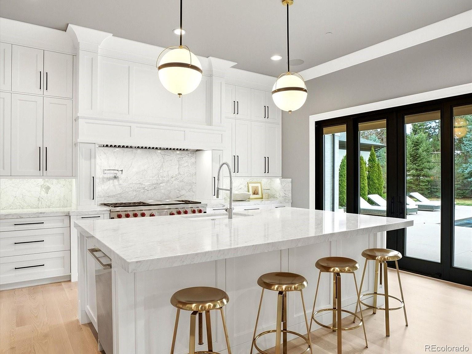 This bright and airy kitchen features a large central island with a waterfall-edge marble countertop and three metallic bar stools. The space is anchored by white cabinetry, a professional-grade range, and elegant globe pendant lighting, all set against a backdrop of marble backsplash and light wood flooring. Large black-framed glass doors provide a seamless transition to the outdoor patio, creating a sophisticated and open-concept living environment.