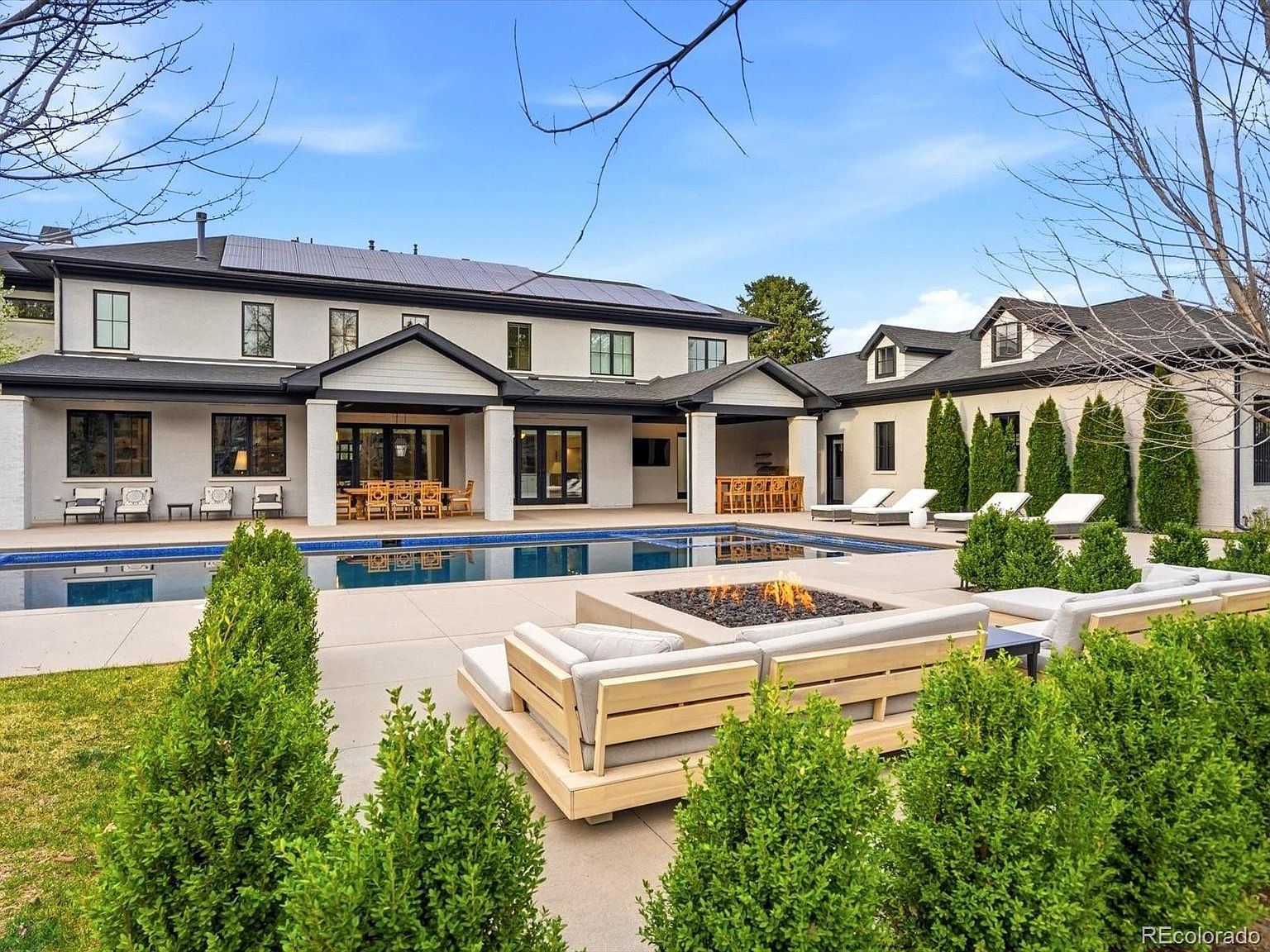 This expansive rear exterior view showcases a luxurious modern estate featuring a large, pristine swimming pool and a central fire pit lounge area. The home boasts a sophisticated design with clean lines, dark-framed windows, and a covered patio space perfect for outdoor dining and entertaining. The meticulously landscaped grounds, including manicured hedges and a spacious concrete deck, create an inviting and high-end atmosphere.