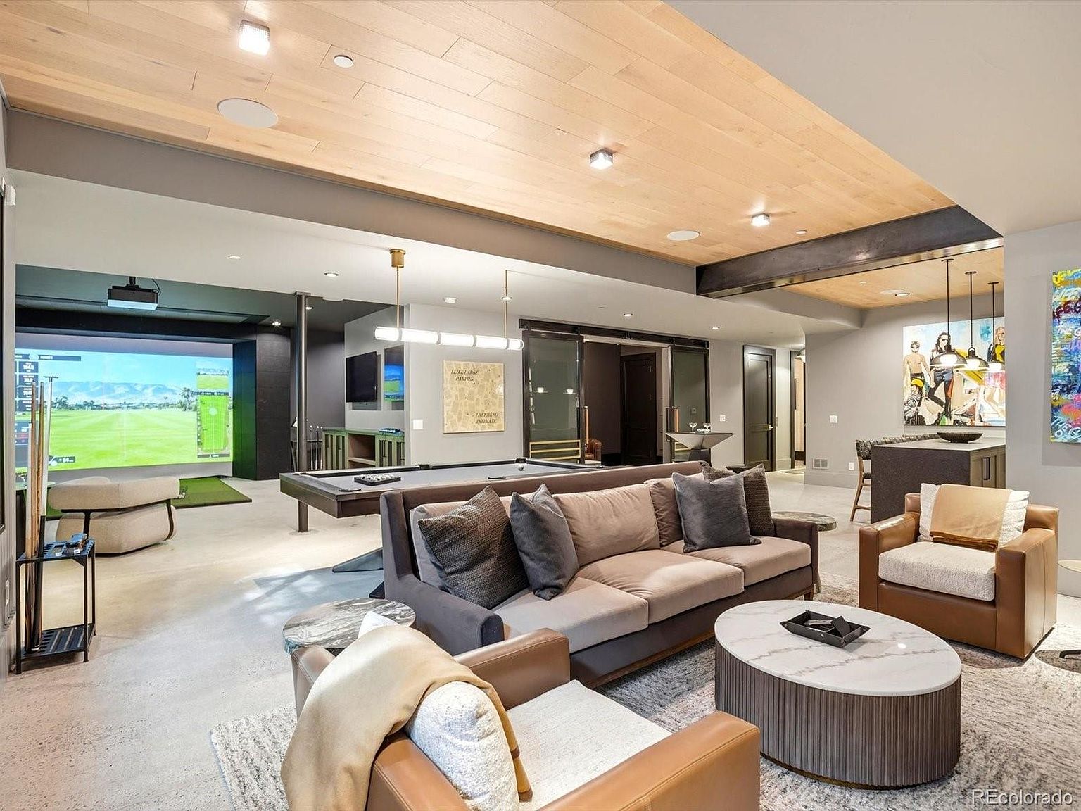 This expansive, modern basement serves as a high-end entertainment hub, featuring a golf simulator, a professional-grade pool table, and a comfortable lounge area with plush seating. The space is defined by a striking wood-paneled ceiling, recessed lighting, and a sophisticated neutral color palette that creates a luxurious yet inviting atmosphere. The open-concept layout seamlessly integrates various recreational zones, making it an ideal space for hosting and leisure.