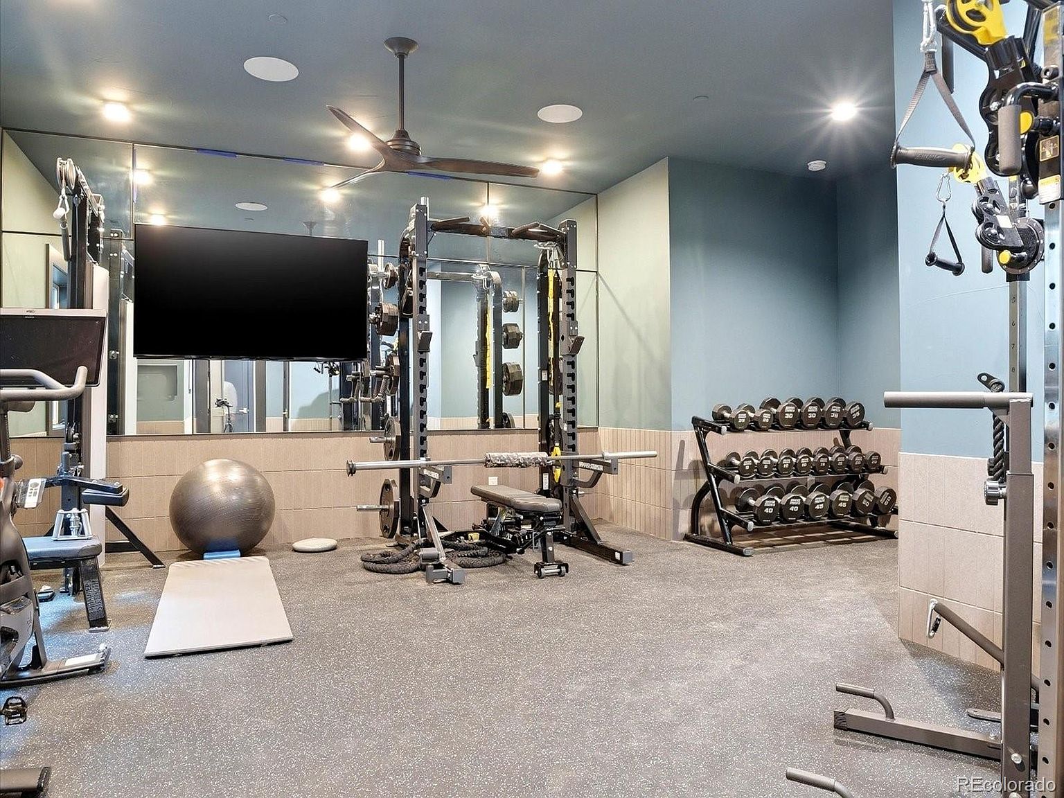 This well-equipped home gym features a professional-grade power rack, a comprehensive dumbbell rack, and a stability ball, all set against a mirrored wall that enhances the sense of space. The room is finished with durable, dark-speckled rubber flooring and a modern ceiling fan, creating a functional and inviting environment for fitness. A large wall-mounted television provides entertainment, making this a complete and versatile workout area.