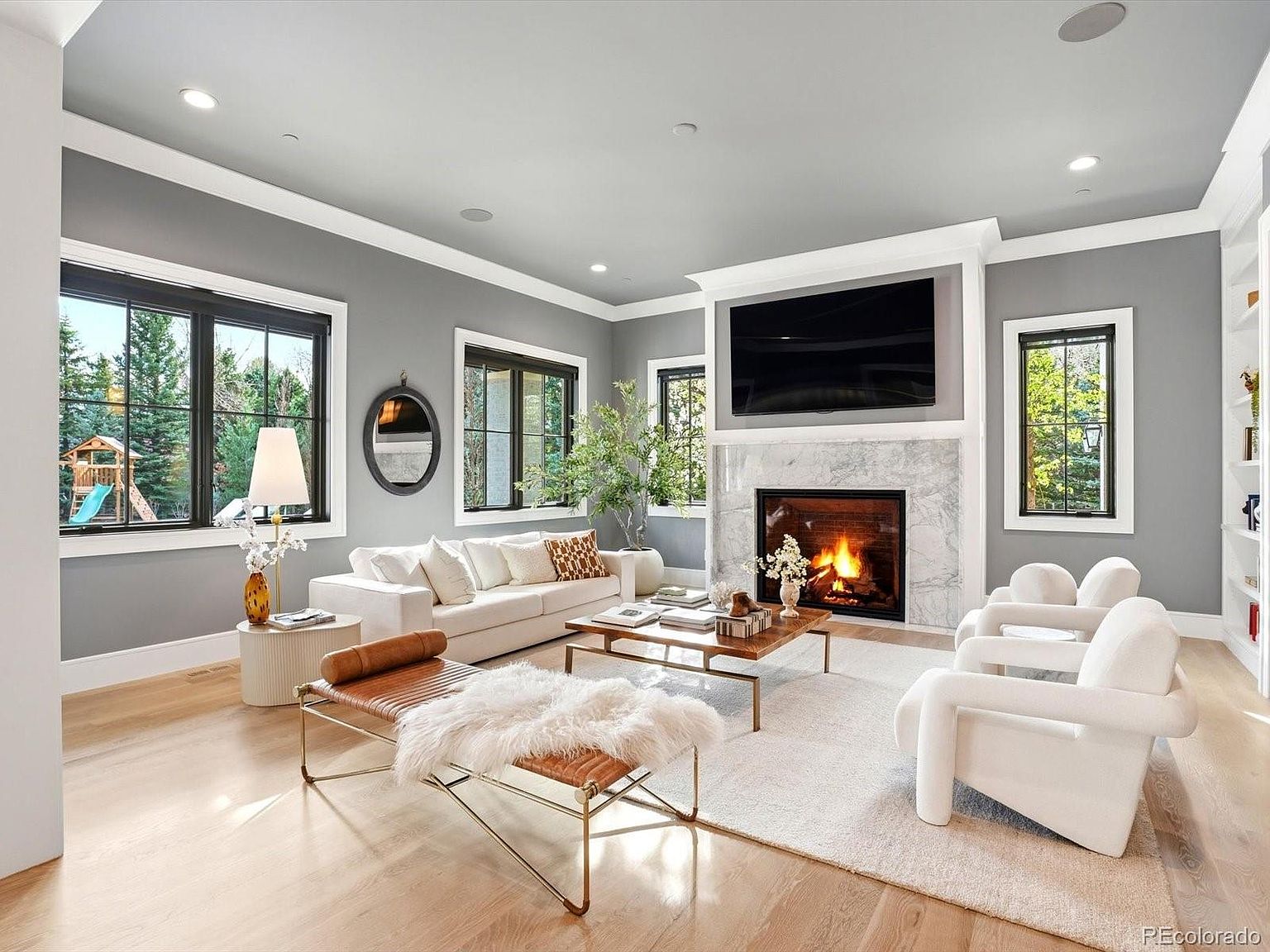 This elegant living room features a sophisticated gray and white color palette, centered around a marble-surround fireplace with an integrated flat-screen television. The space is furnished with a plush white sofa, two modern sculptural armchairs, and a leather bench with a faux-fur throw, all arranged on a soft area rug over light hardwood floors. Large windows with black frames provide ample natural light and views of the backyard, creating a bright, inviting, and high-end atmosphere.