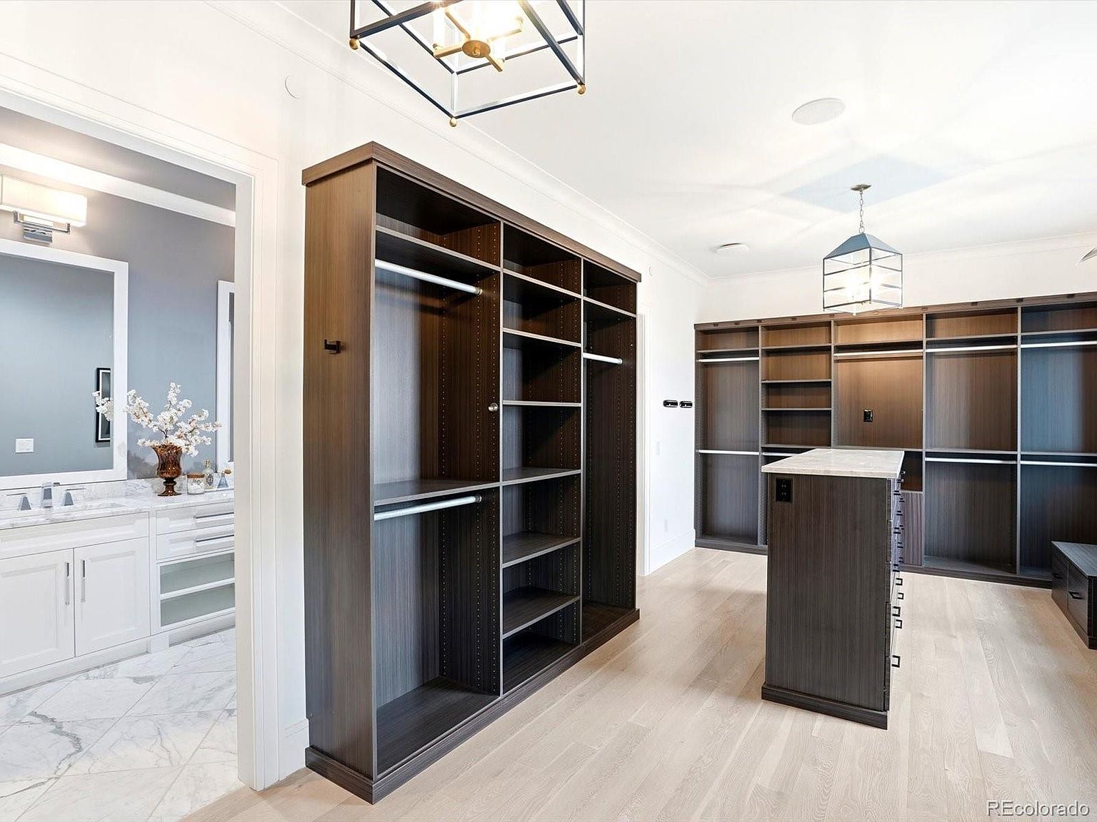 This spacious, high-end walk-in closet features dark wood cabinetry with extensive shelving and hanging space, centered around a functional island with a stone countertop. The room is bright and airy, with light hardwood flooring and elegant pendant lighting, while a doorway leads directly into a luxurious, marble-tiled bathroom. The overall aesthetic is sophisticated, organized, and designed for a premium lifestyle.