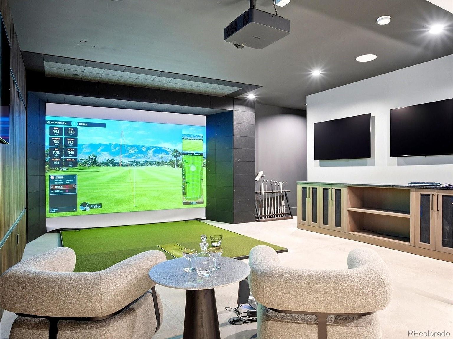 This image showcases a high-end home golf simulator room, featuring a large projection screen displaying a virtual course, a putting green mat, and a rack of golf clubs. The space is furnished with two modern, curved armchairs and a small marble-topped table, while two wall-mounted televisions provide additional entertainment options. The room is designed with a sophisticated, contemporary aesthetic, utilizing recessed lighting and a neutral color palette to create an inviting atmosphere for leisure and practice.