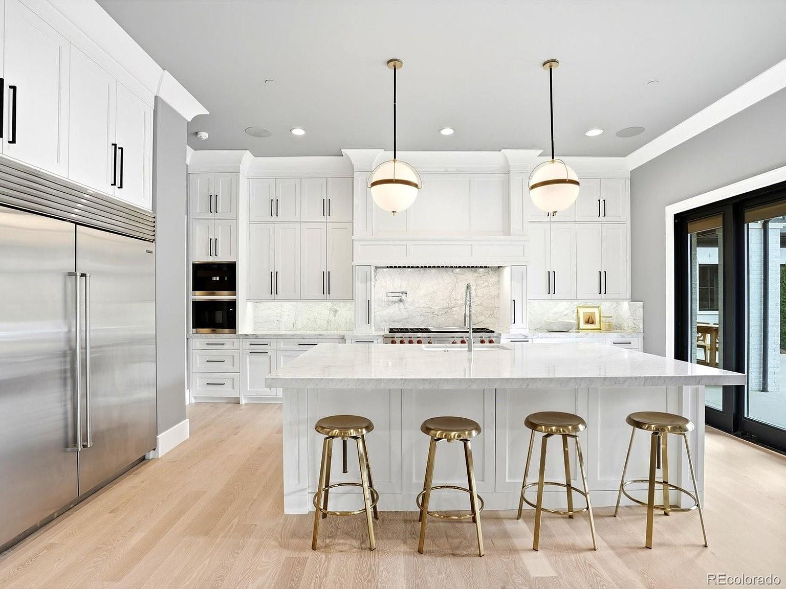 This bright and airy kitchen features a large central island with a marble countertop and four metallic bar stools, serving as the focal point of the room. The space is equipped with high-end stainless steel appliances, crisp white cabinetry, and elegant globe pendant lighting that adds a touch of sophistication. The light hardwood flooring and neutral gray walls create a clean, modern aesthetic that feels both luxurious and inviting.