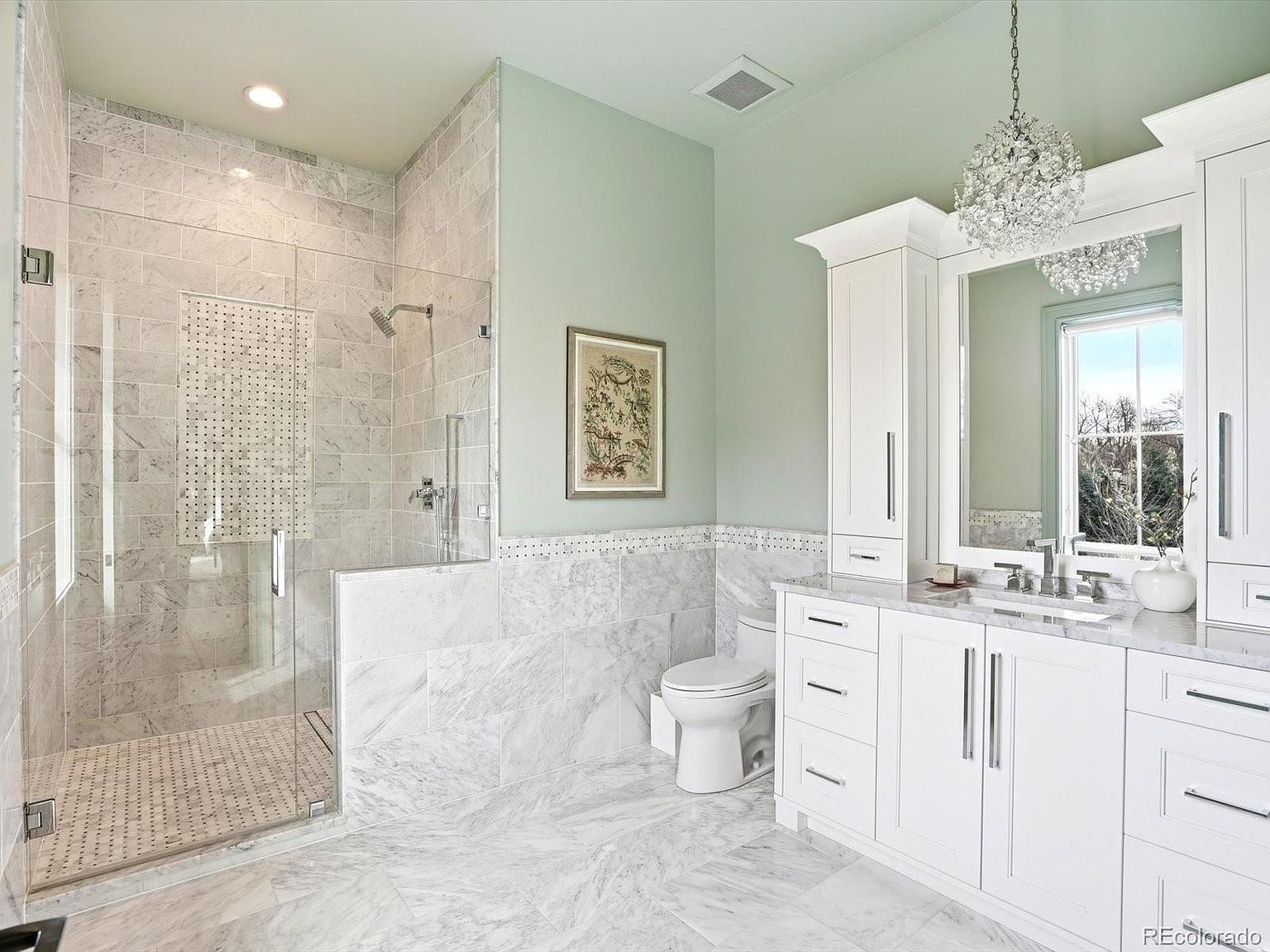 This elegant primary bathroom features a luxurious walk-in shower with marble tile walls and a glass enclosure, paired with a crisp white vanity topped with matching marble. The room is painted in a soft sage green, complemented by a sparkling crystal chandelier that adds a touch of sophistication. The bright, clean aesthetic and high-end finishes create a serene and spa-like atmosphere.