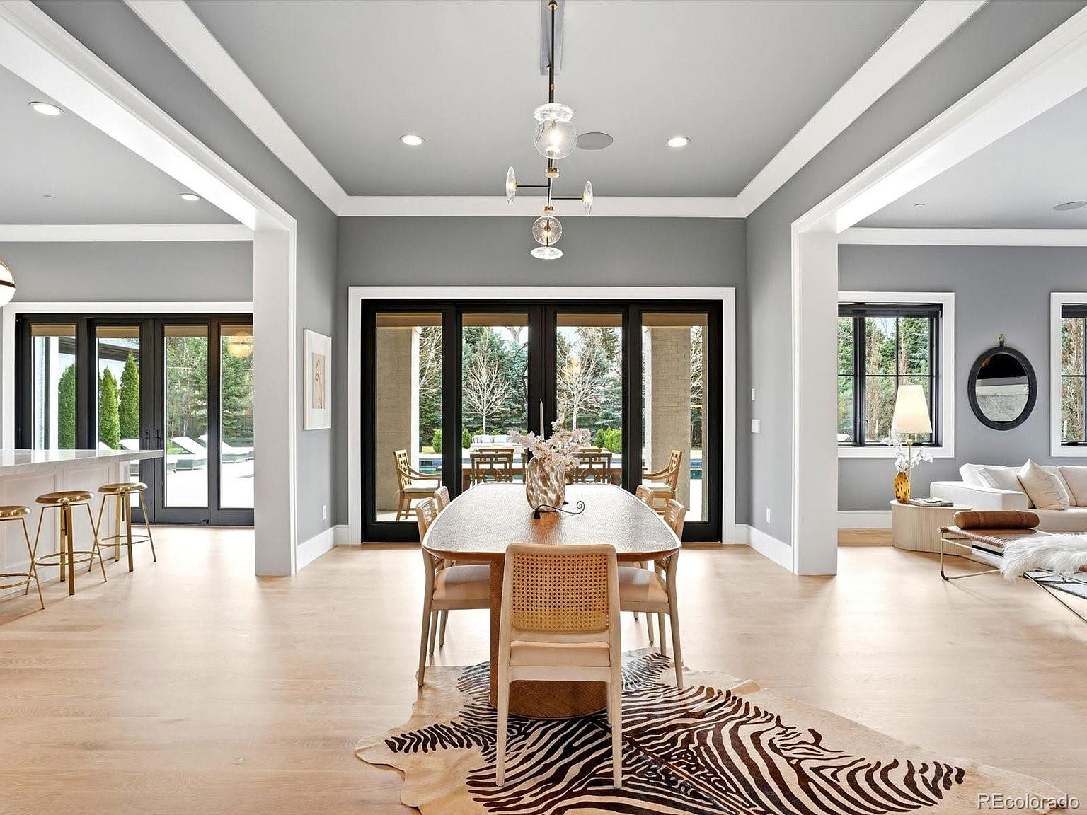 This elegant dining room features a contemporary aesthetic with a central wooden table surrounded by light-toned cane-back chairs, all set atop a striking zebra-print area rug. The space is framed by high ceilings with crown molding, a modern glass chandelier, and expansive black-framed glass doors that provide a seamless transition to the outdoor patio. The neutral color palette and open-concept layout create a sophisticated and airy atmosphere perfect for formal entertaining.