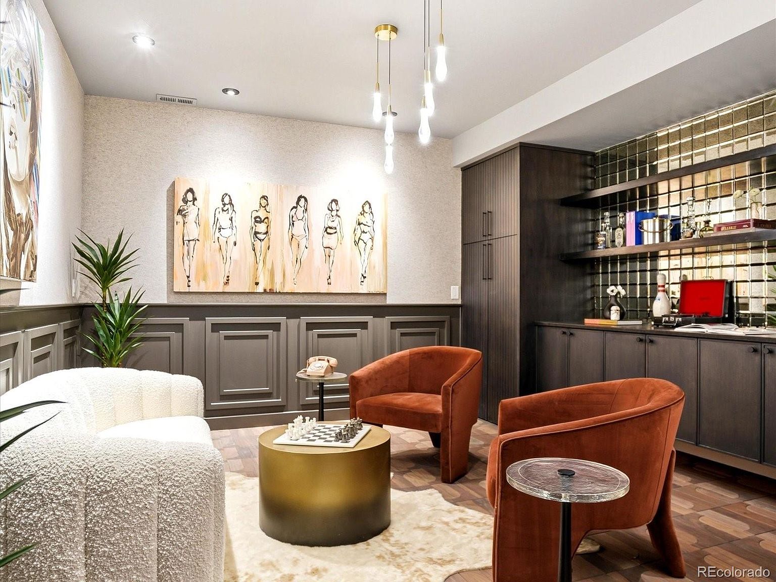 This sophisticated lounge area features a contemporary design with a rich color palette, highlighted by two burnt-orange velvet armchairs and a textured white sofa. The room is anchored by a gold cylindrical coffee table topped with a chessboard, set against a backdrop of dark wood paneling and a mirrored bar wall. A large, stylized figurative art piece serves as a focal point, while modern pendant lighting adds a touch of elegance to this stylish, intimate space.