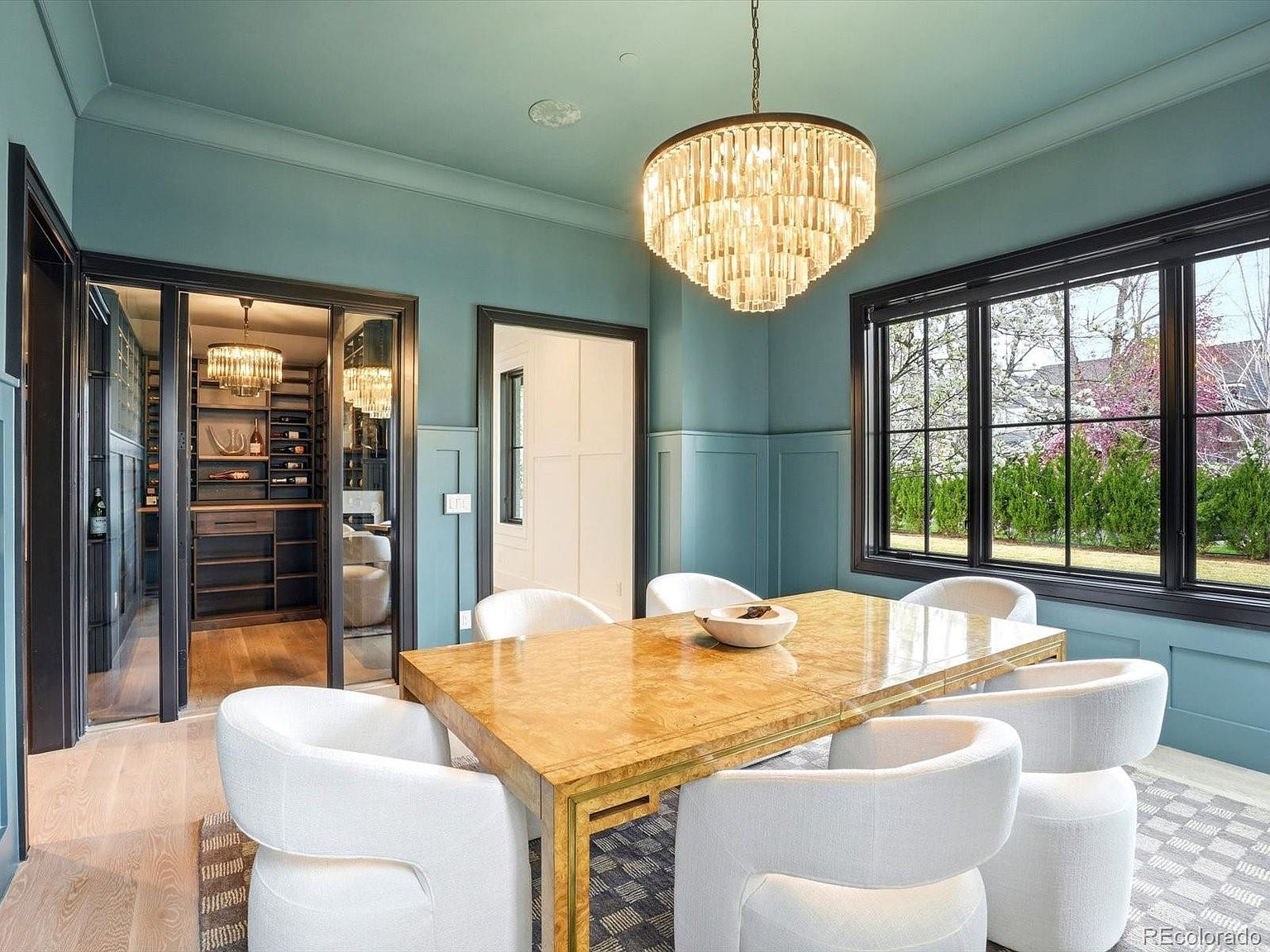 This elegant dining room features a striking burl wood table surrounded by six modern, cream-colored upholstered chairs, all set beneath a grand, tiered crystal chandelier. The walls are painted a sophisticated teal and accented with dark trim, while a large window provides a view of the lush greenery outside. A glass-paneled doorway leads into a custom-built wine cellar, adding a touch of luxury and depth to the space.