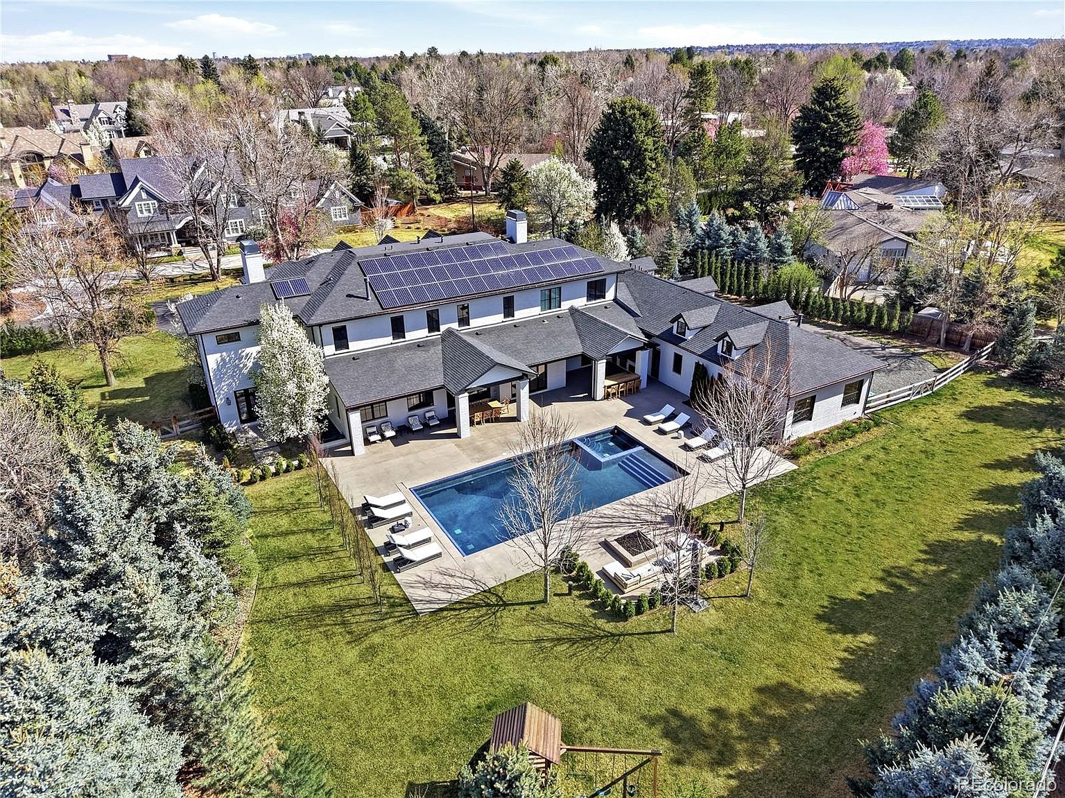 This high-angle aerial view captures a luxurious, modern estate featuring a sprawling residence with a dark shingled roof equipped with solar panels. The property boasts a large, pristine swimming pool and integrated spa, surrounded by a spacious concrete patio, a fire pit area, and meticulously landscaped green lawns. The setting is serene and upscale, nestled within a lush, tree-lined neighborhood that suggests a high-end suburban lifestyle.