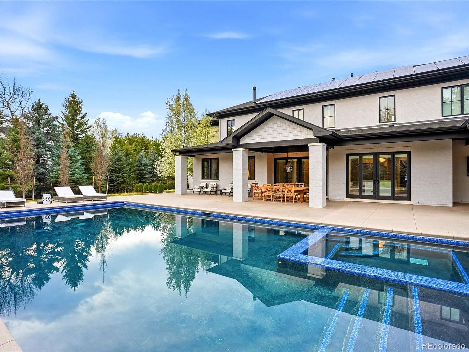 This stunning rear exterior view showcases a luxurious swimming pool and integrated spa, set against a modern, light-colored home with a covered patio area. The expansive concrete deck features outdoor dining and lounge furniture, perfect for entertaining, while the surrounding mature trees provide a serene and private backdrop. The scene is captured from a low, cinematic angle that emphasizes the inviting blue water and the seamless transition between indoor and outdoor living spaces.