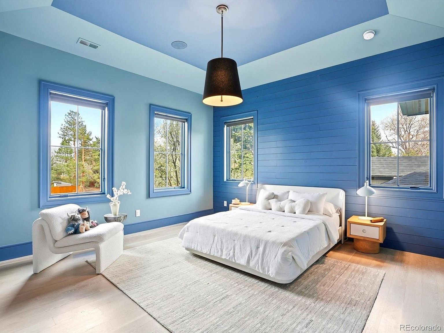 This spacious primary bedroom features a striking blue color palette with a shiplap accent wall behind a modern white bed. The room is bathed in natural light from multiple large windows, complemented by a contemporary black pendant light and a plush white armchair. The light wood flooring and neutral area rug create a serene, sophisticated atmosphere perfect for a master suite.