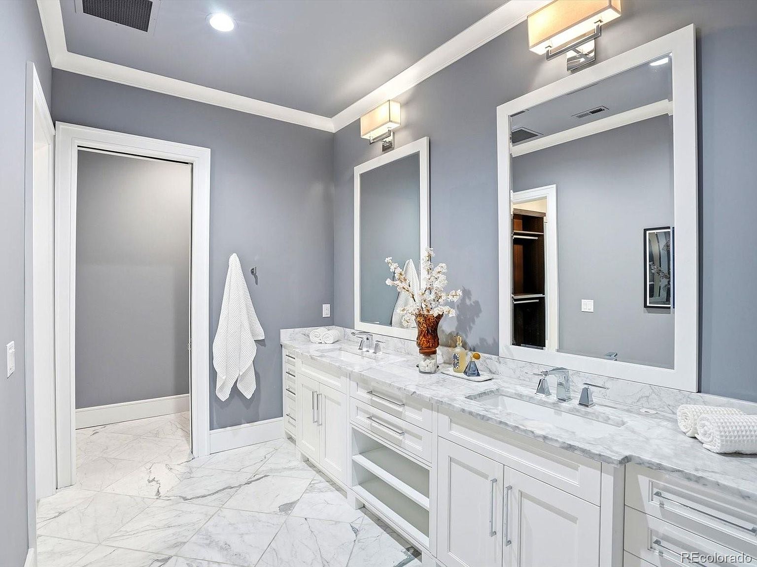 This elegant primary bathroom features a double vanity with white cabinetry, marble countertops, and dual rectangular mirrors illuminated by modern wall sconces. The space is finished with light gray walls, white trim, and marble-patterned tile flooring, creating a clean and sophisticated aesthetic. A doorway leads into a walk-in closet, enhancing the functional layout of this bright and airy room.