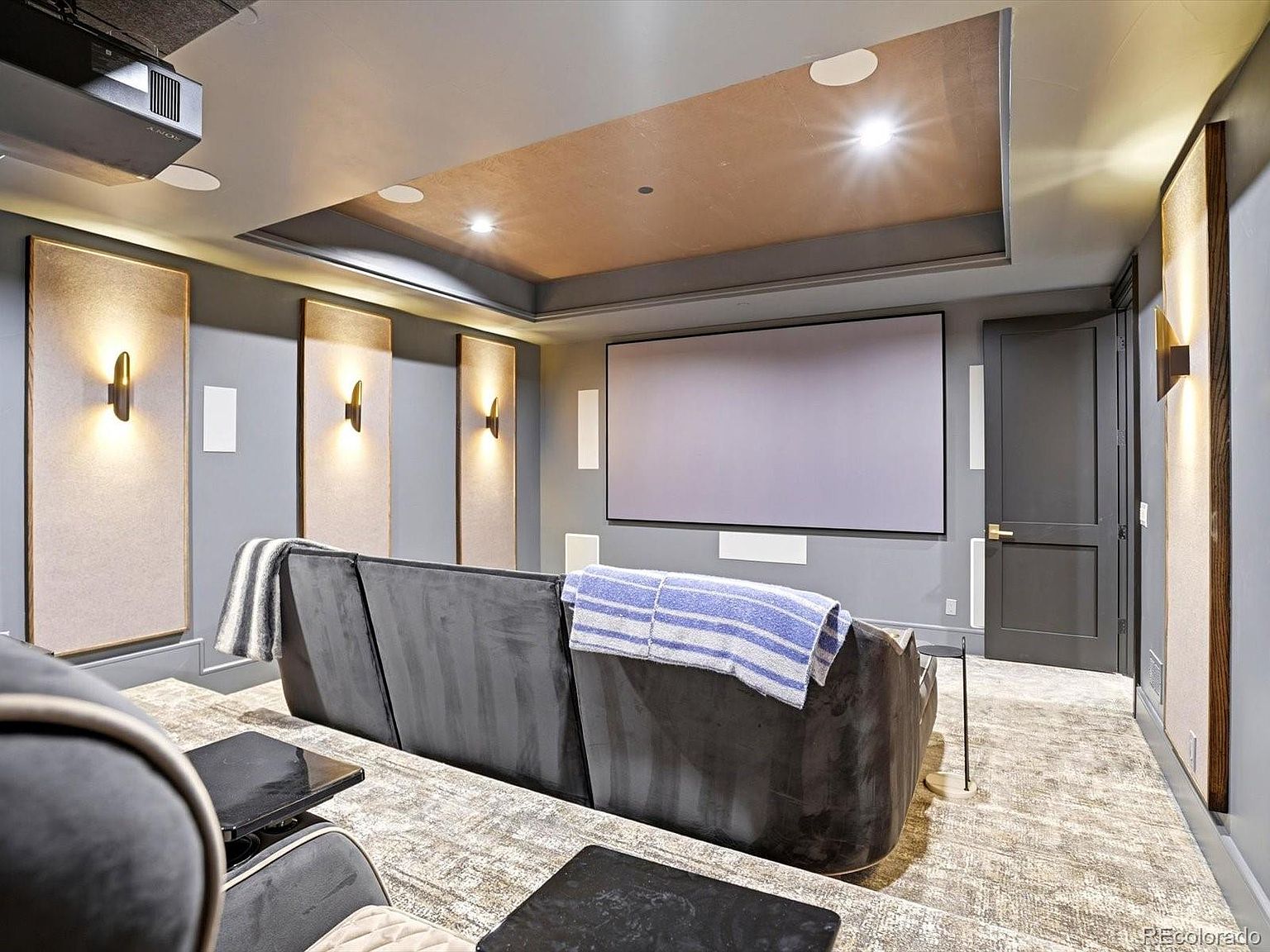 This image showcases a dedicated home theater room featuring a large projection screen, tiered seating with plush dark gray sofas, and ambient wall sconce lighting. The room is designed with acoustic wall panels and a recessed tray ceiling, creating an immersive cinematic environment. The perspective is from the back of the room, looking toward the screen, emphasizing the comfortable and professional layout of the space.