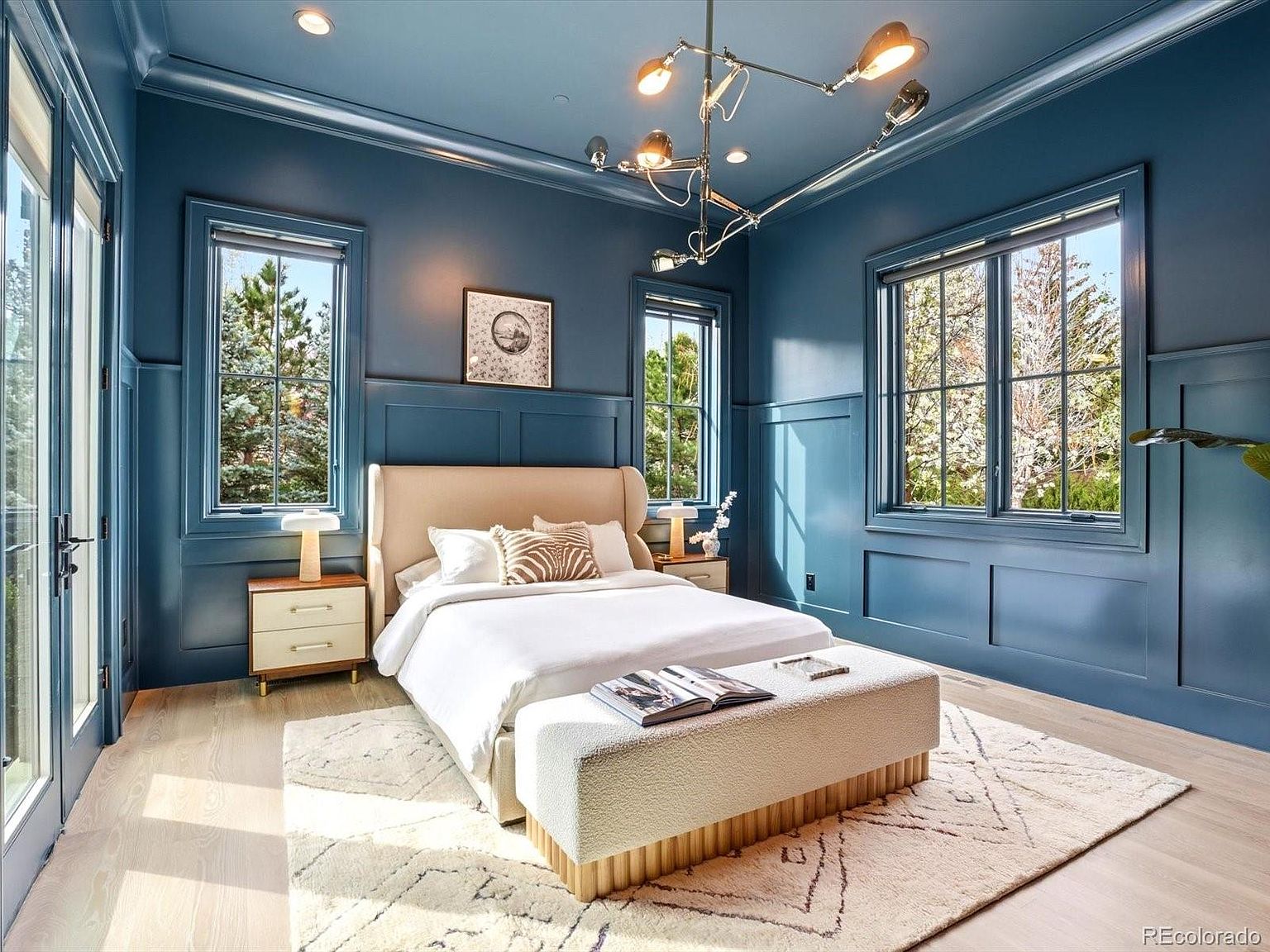 This elegant primary bedroom features deep blue wainscoting that creates a sophisticated, moody atmosphere, contrasted by light hardwood floors and a neutral-toned bed. A modern, multi-armed chandelier serves as a striking focal point, while large windows allow ample natural light to fill the space. The room is furnished with a plush bench at the foot of the bed and mid-century modern nightstands, offering a blend of contemporary style and comfort.