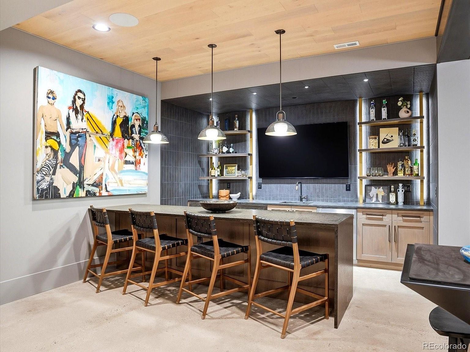 This modern basement bar area features a sleek, dark-tiled wet bar with integrated shelving, a mounted television, and a spacious countertop with four woven-seat bar stools. The space is accented by a large, vibrant pop-art painting on the left wall and a warm wood-paneled ceiling, creating a sophisticated yet relaxed entertainment atmosphere. The perspective is a wide, eye-level shot that captures the full functionality and stylish design of the home lounge.