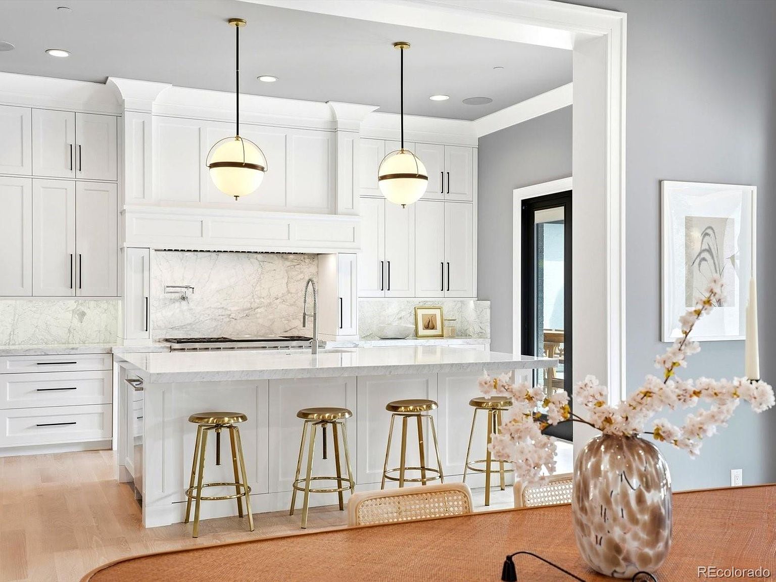 This elegant, high-end kitchen features crisp white cabinetry, marble countertops, and a spacious central island with four gold-toned bar stools. Two sophisticated globe pendant lights hang above the island, complementing the bright, airy aesthetic and the marble backsplash. The perspective is captured from a dining area, offering a clear view of the kitchen's clean lines and luxurious finishes.