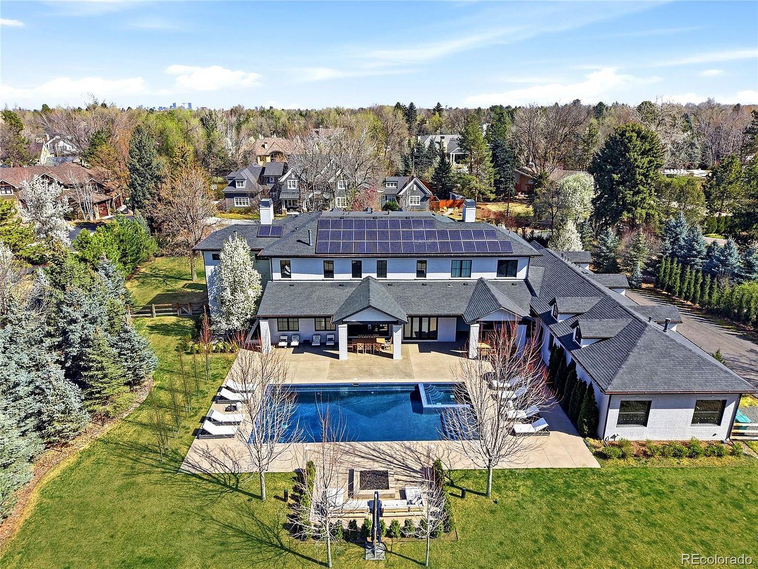 This stunning aerial view captures a luxurious multi-story residence featuring a dark shingled roof equipped with solar panels and a crisp, white exterior. The property boasts an expansive backyard complete with a pristine rectangular swimming pool, a sunken fire pit lounge area, and manicured landscaping. The perspective highlights the home's impressive scale and its seamless integration into a lush, tree-lined suburban neighborhood.