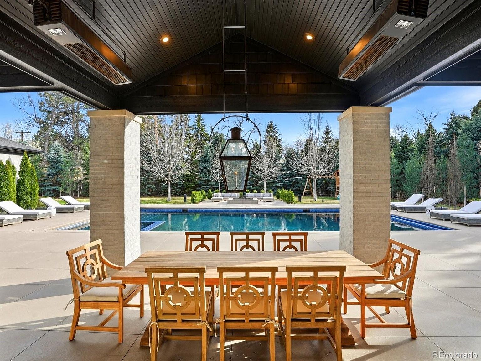 This luxurious covered outdoor dining area features a large wooden table with matching lattice-back chairs, set beneath a vaulted, dark-stained wood ceiling with integrated heaters. The space opens directly onto a pristine swimming pool and a landscaped backyard, creating a seamless transition between indoor comfort and outdoor leisure. The symmetrical composition and elegant lantern lighting evoke a sophisticated, resort-style atmosphere perfect for entertaining.