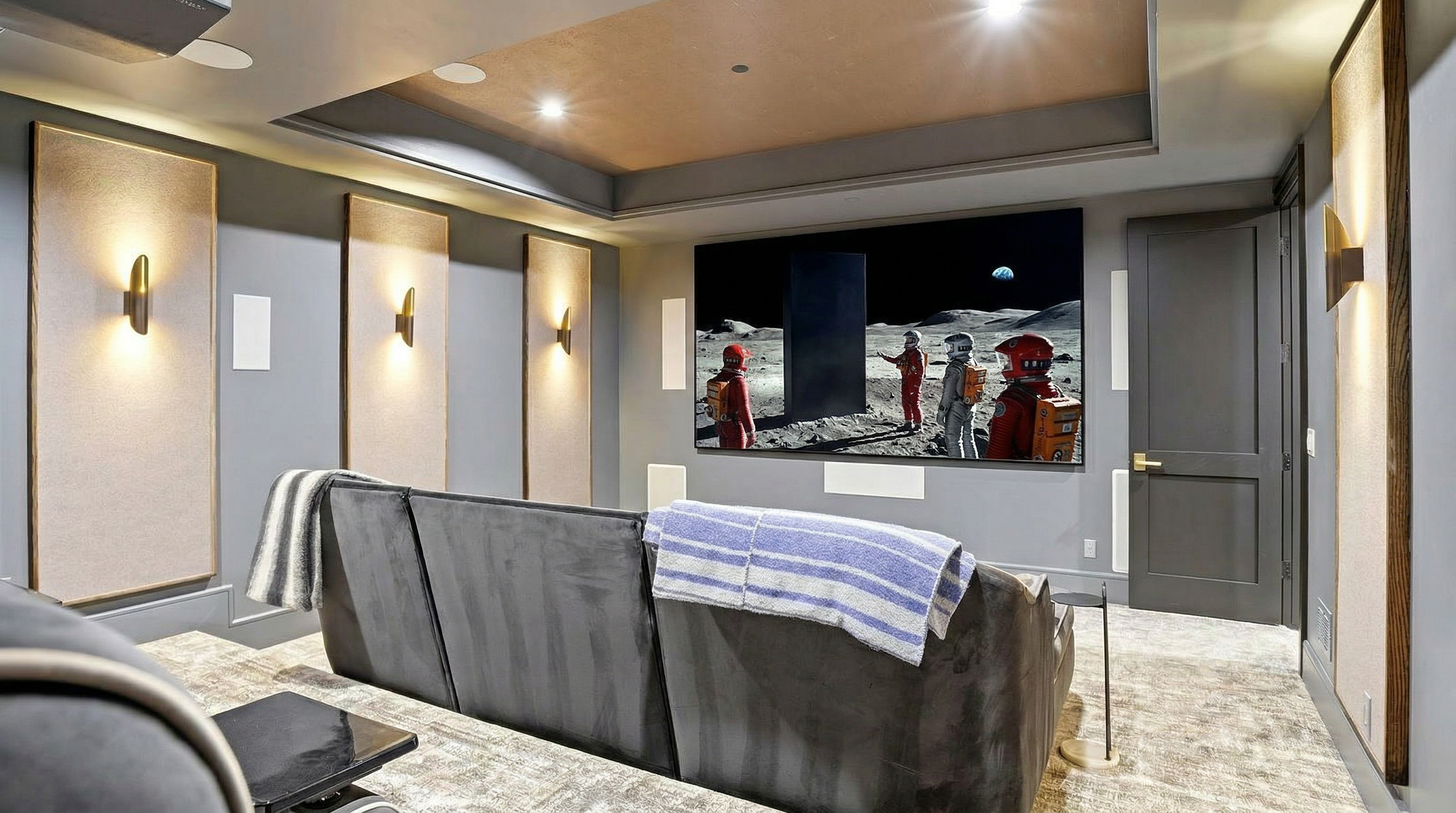 This dedicated home theater room serves as an impressive entertainment space, furnished with comfortable reclining lounge seating and a large projection screen displaying a classic film scene. The room features elegant wall sconces, integrated sound-dampening acoustic wall panels with wood trim, and a tray ceiling with recessed lighting. The overall design emphasizes a luxurious and immersive cinematic experience.