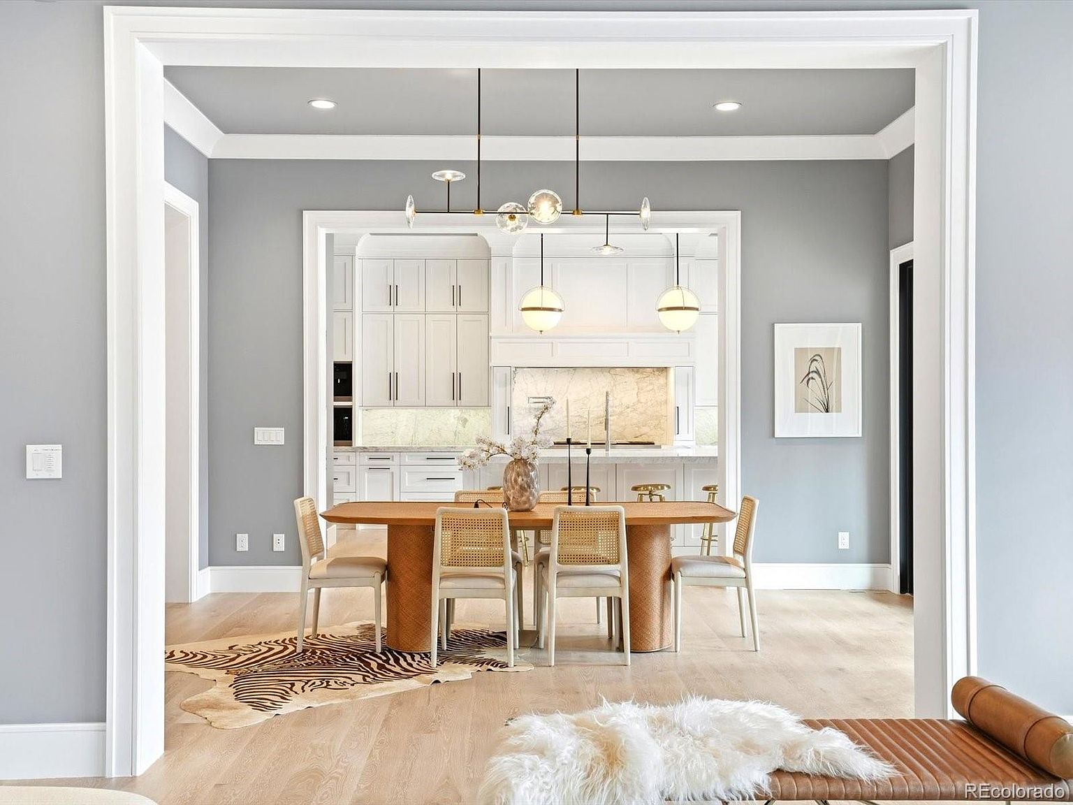 This elegant dining room features a modern wooden table paired with stylish cane-back chairs, set atop a distinctive zebra-print area rug. The space is framed by a wide, white-trimmed doorway that looks directly into a bright, high-end kitchen with marble backsplashes and white cabinetry. A sophisticated multi-globe chandelier hangs above the table, creating a warm and inviting atmosphere that blends contemporary design with classic textures.