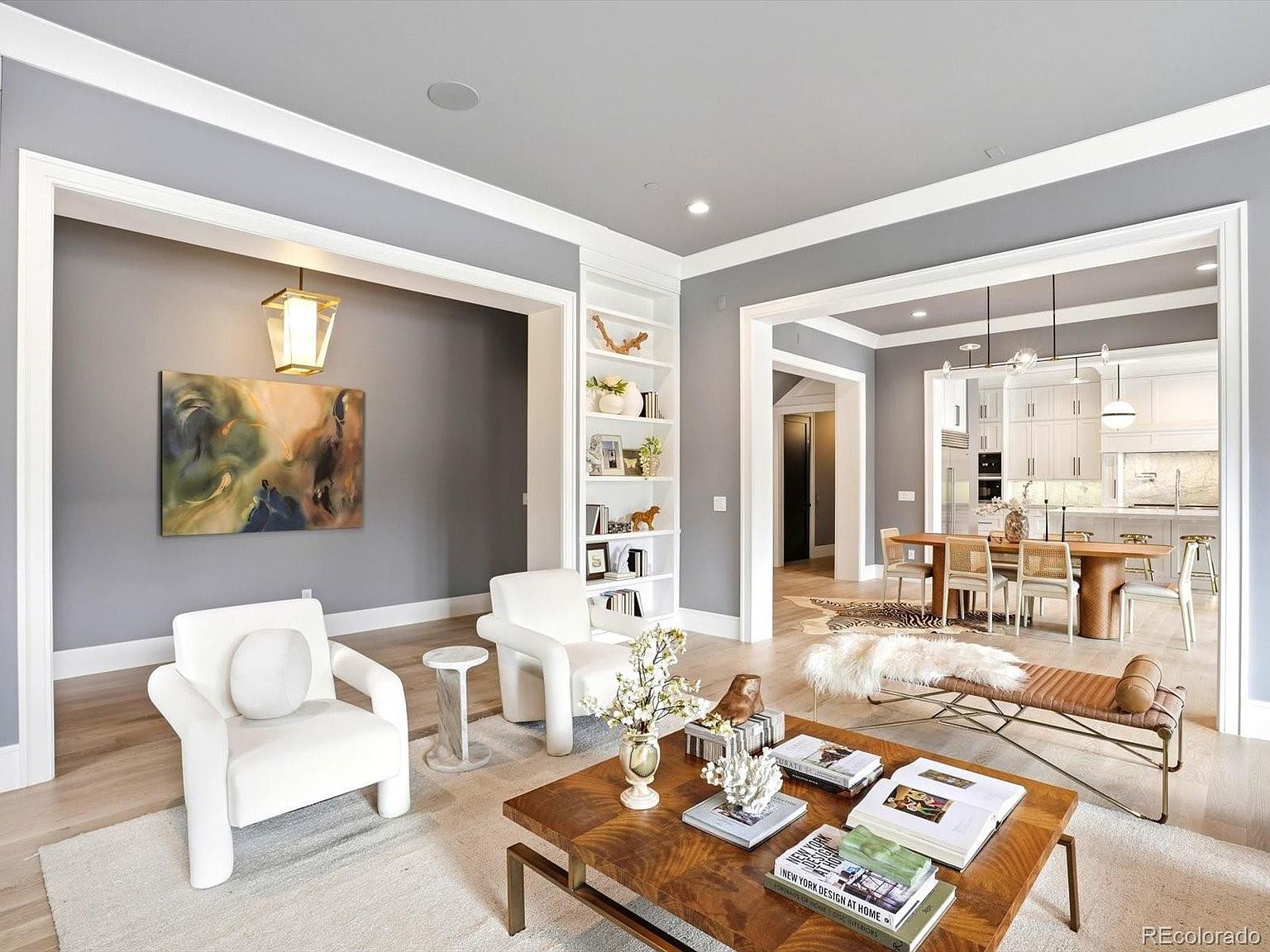 This elegant living room features a sophisticated neutral color palette with plush white armchairs, a rich wooden coffee table, and a sleek leather bench. The space is framed by architectural archways that lead into a bright, open-concept dining area and kitchen, creating a seamless flow throughout the home. A built-in bookshelf and curated decor add a refined, high-end touch to this inviting and well-lit living space.