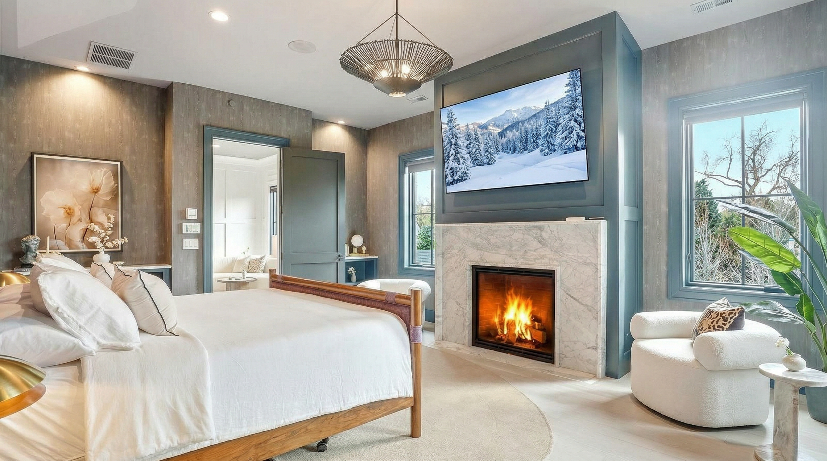 This luxurious primary bedroom features a sophisticated design with light gray wood-look paneling and a striking blue accent wall surrounding a white marble fireplace. The room includes a spacious bed, a cozy white armchair, a decorative light fixture, and a flat-screen TV, creating a warm and refined atmosphere. Large windows and an open doorway provide plenty of natural light, emphasizing the high-end, comfortable feel of the space.