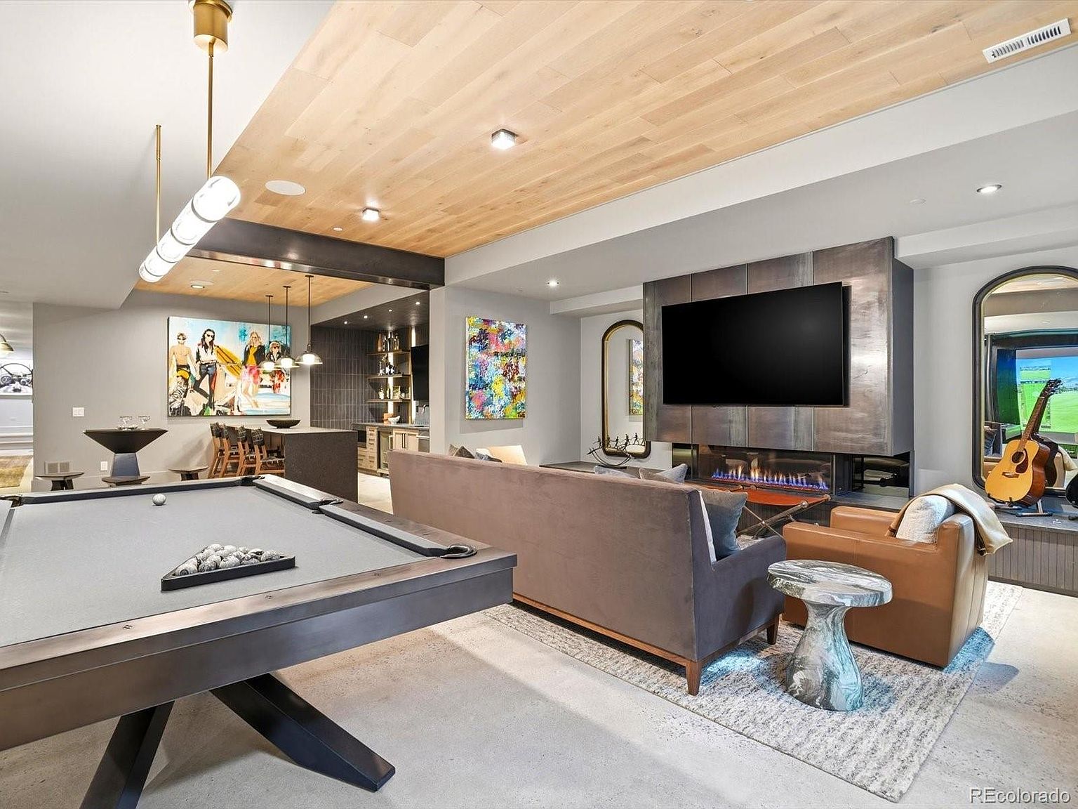This sophisticated basement living area features a modern pool table in the foreground and a cozy lounge space with a fireplace and television. The room is accented by a warm wood-paneled ceiling, industrial-style metal finishes, and vibrant contemporary artwork, creating a high-end entertainment environment. The perspective captures the open-concept layout, seamlessly connecting the game area to the bar and seating zones.