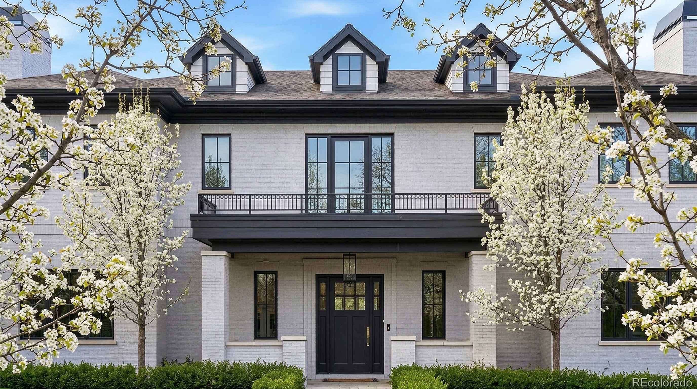 This elegant, symmetrical two-story home features a sophisticated white brick facade complemented by bold black window frames, dark trim, and a matching black front door. The exterior is enhanced by three charming gabled dormers on the roof and a prominent balcony above the main entryway, all framed by mature, blossoming white trees that add to its curb appeal.