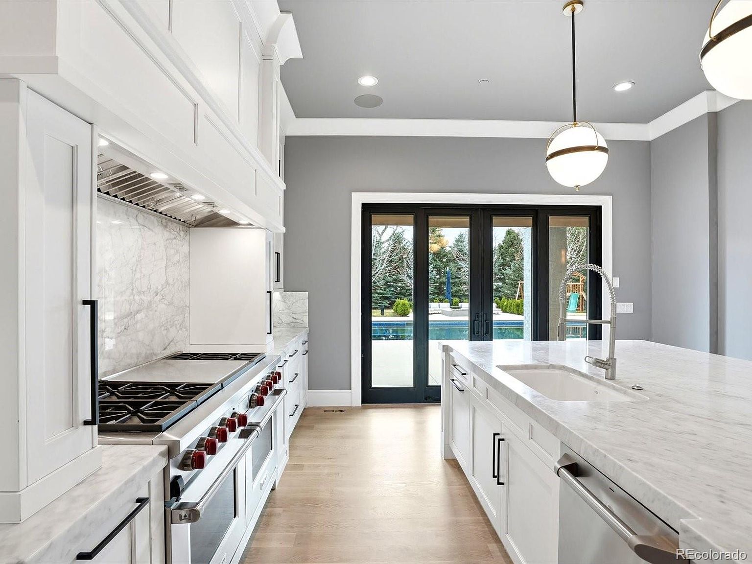 This bright and airy kitchen features a large marble-topped island, professional-grade white range, and custom cabinetry, creating a sophisticated and clean aesthetic. The space is anchored by a striking pendant light and opens through expansive black-framed glass doors to a view of a backyard pool. The combination of light wood flooring, marble countertops, and gray walls provides a modern, high-end feel perfect for luxury living.