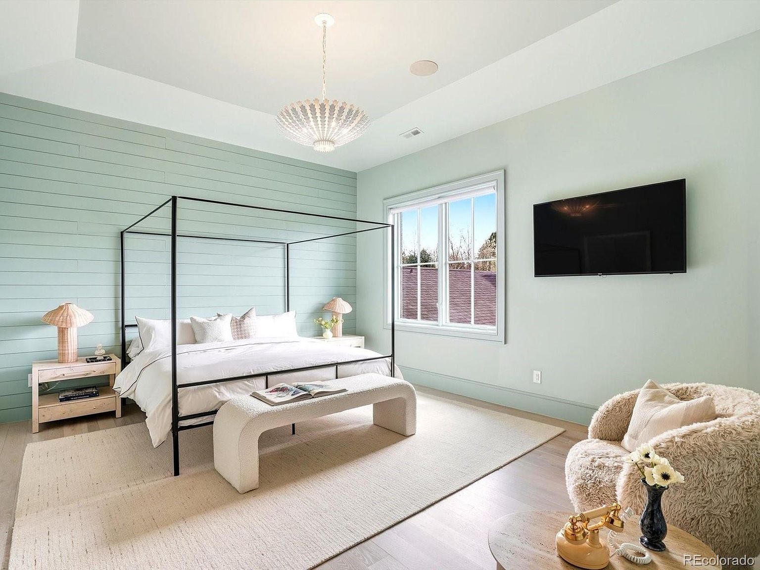 This serene primary bedroom features a striking black metal canopy bed set against a light sage green shiplap accent wall. The room is furnished with a textured cream-colored bench, a plush fuzzy armchair, and light wood nightstands, creating a soft, modern aesthetic. A large window allows natural light to fill the space, while a central chandelier adds a touch of elegance to the airy, high-ceilinged room.