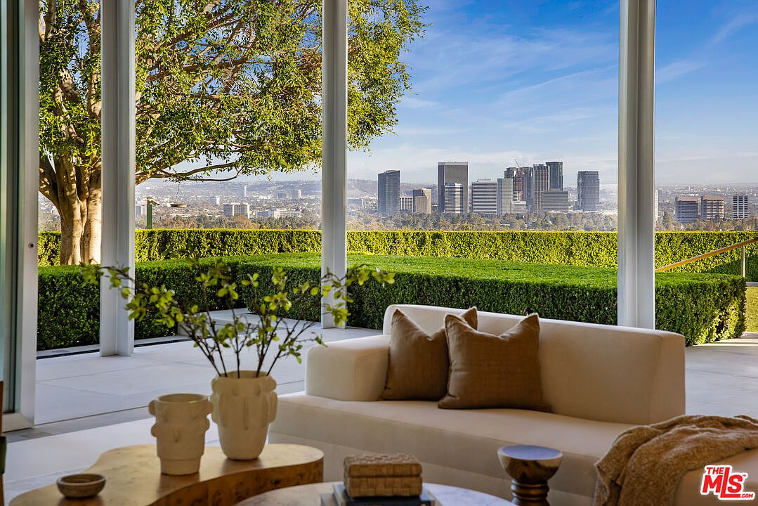 This image showcases a luxurious living room with a stunning view of a cityscape through large glass windows. The interior features a modern white sofa with brown pillows, complemented by stylish coffee tables and decorative vases. The room exudes elegance and sophistication, emphasizing the seamless integration of indoor and outdoor spaces.