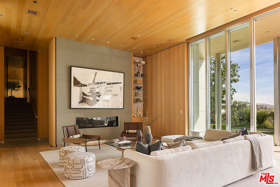 This is an interior shot of a modern living room featuring a large abstract painting above a fireplace. The room is well-lit with natural light from large windows, and the decor includes a neutral-colored sofa, two leather chairs, and patterned ottomans. The space is designed with clean lines and a minimalist aesthetic, creating a sophisticated and inviting atmosphere.