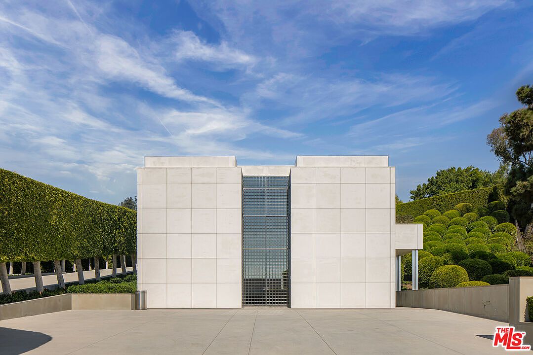 This is a front exterior view of a modern, minimalist building with a grid-like facade. The building features a central glass section flanked by solid, tiled blocks. The surrounding landscape includes manicured hedges and trees, creating a clean and structured environment. The perspective is a straight-on shot, emphasizing the symmetry and geometric design of the building.