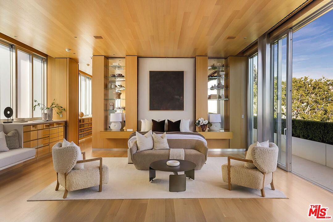 This is a luxurious primary bedroom featuring a light wood floor and ceiling, a large bed with a neutral color palette, and two plush armchairs flanking a round coffee table. Built-in shelving units with glass displays add a touch of elegance, while large sliding glass doors offer access to a balcony and views of the outdoors. The room exudes a sense of calm and sophistication.