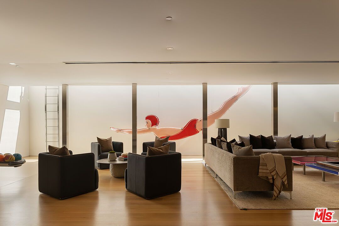 This is an interior shot of a modern living room featuring hardwood floors, a neutral color palette, and contemporary furnishings. The room is well-lit, with natural light filtering through large windows adorned with a unique swimmer art piece. The space includes a sectional sofa, armchairs, and a coffee table, creating a comfortable and stylish living area.