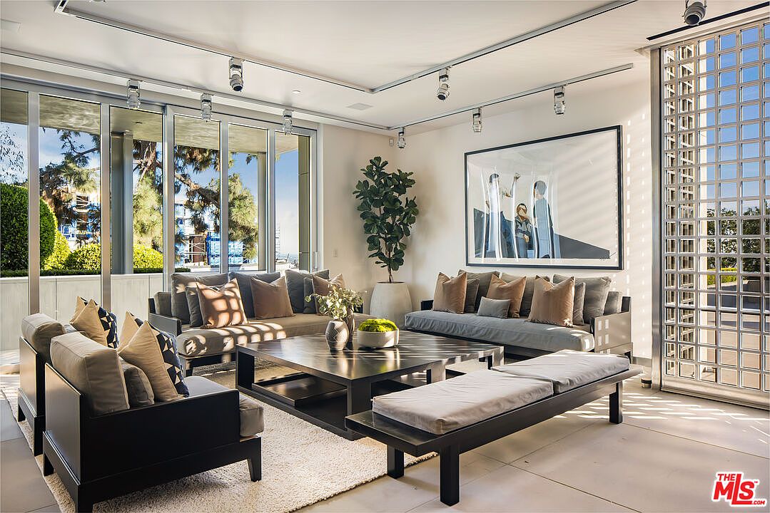 This is a bright and modern living room featuring contemporary furniture, including sofas, chairs, and a coffee table. Large windows provide ample natural light and views of the outdoors, while track lighting illuminates the space. The room is decorated with neutral tones and artwork, creating a sophisticated and inviting atmosphere.