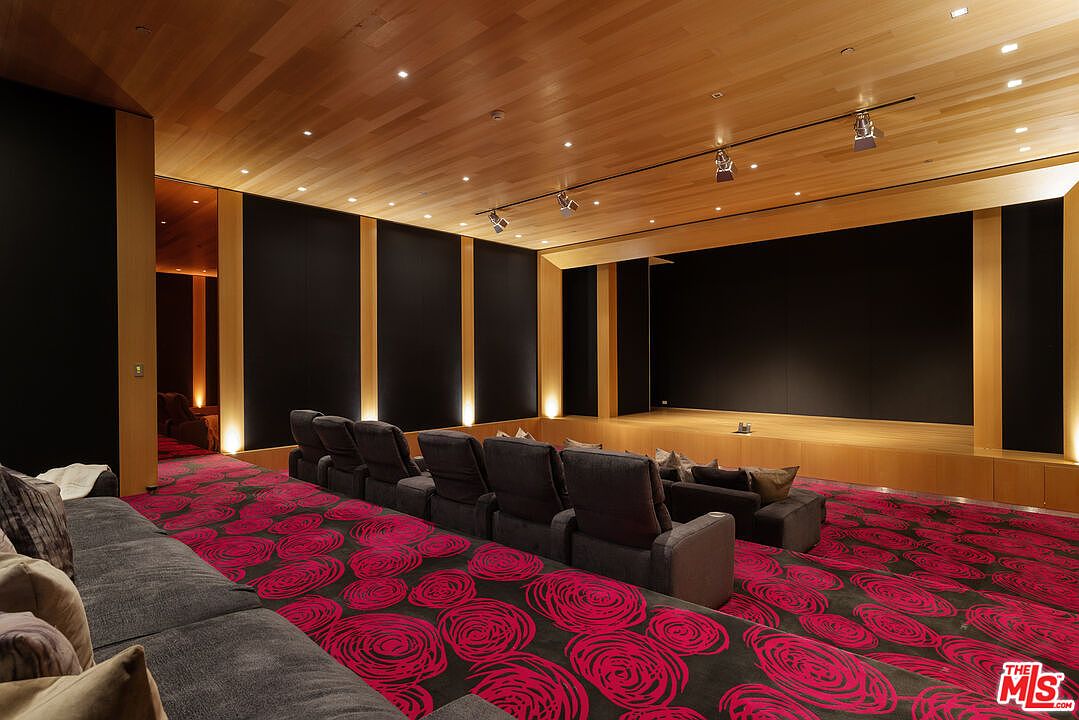 This is a luxurious home theater featuring tiered seating with plush, gray reclining chairs and a large, dark screen framed by wood paneling. The ceiling is finished with light wood planks and recessed lighting, while the floor is covered in a vibrant red and black patterned carpet. The room exudes a sense of comfort and high-end entertainment.