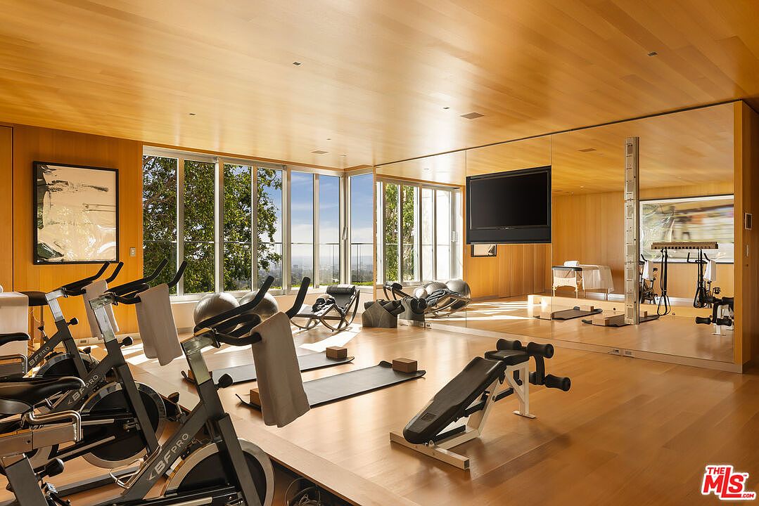 This is a spacious home gym featuring wood floors and wood paneled walls. The room is equipped with exercise bikes, free weights, yoga mats, and a weight bench. Large windows offer natural light and views, while a mirrored wall with a mounted TV enhances the workout experience.